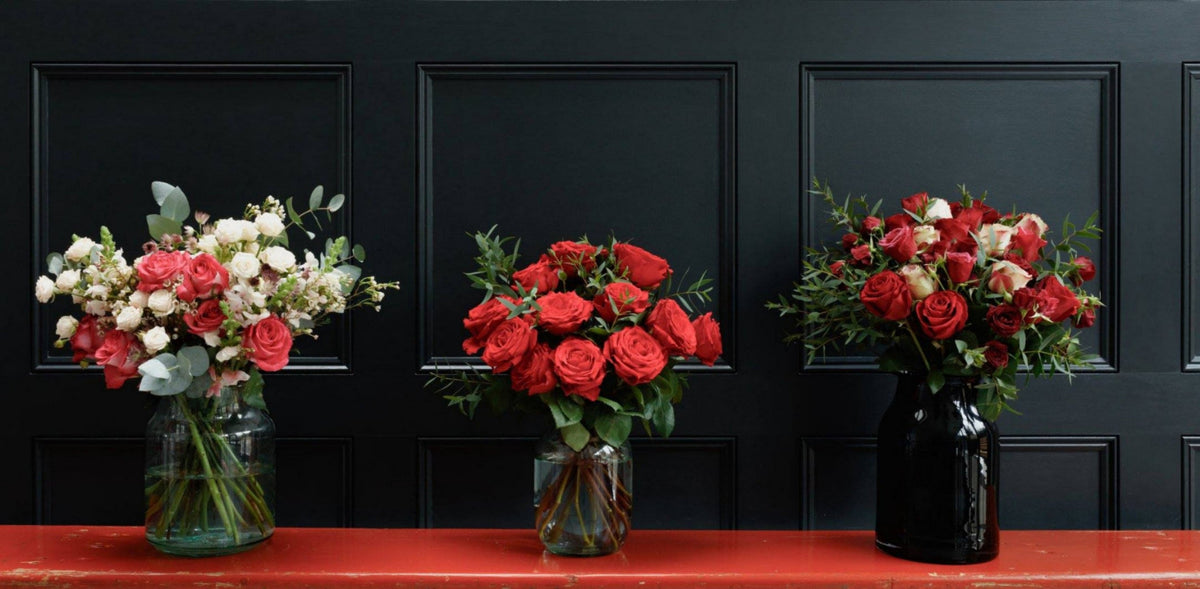 Three floral arrangements in glass vases displayed on a red table against a black paneled wall. The left arrangement contains pink, red, and white flowers with green foliage; the center arrangement features a dense bouquet of red roses; the right arrangement includes red and white flowers with greenery in a glossy black vase.